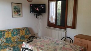 A room with a bed, a table with a patterned tablecloth, a television mounted on the wall, and a window with curtains. - Appartamenti San Teodoro VII (San Teodoro)
