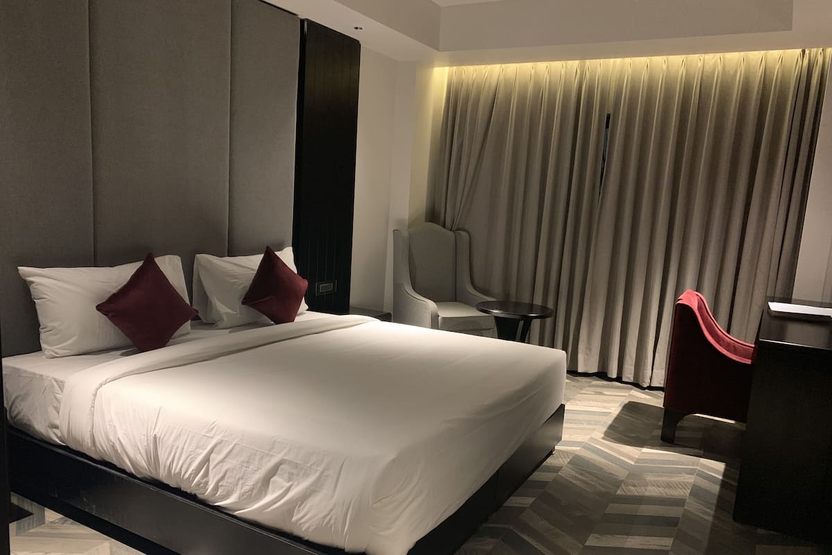 A modern hotel room with a large bed, a desk, a chair, and a view of the city through curtains.