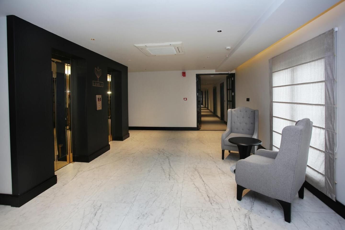 A hotel lobby with a marble floor, a seating area with two armchairs and a small table, and a corridor leading to other rooms.