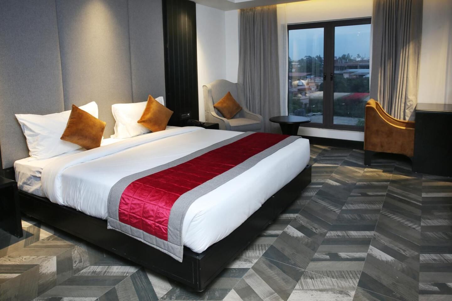 A hotel room with a large bed, two armchairs, a small table, and a view of the city through large windows.