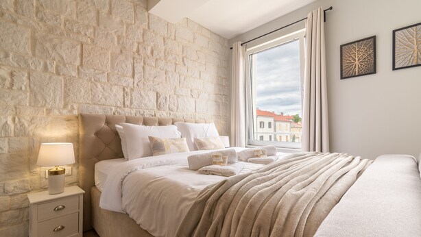 A bedroom with a stone wall, a bed with white linens, a bedside table with a lamp, and a window with curtains. - Luxurious Suites Elegance (Zadar)