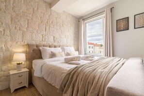 Quarto com parede de pedra, cama com roupa branca, mesa de cabeceira com abajur e janela com cortinas.