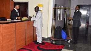 A reception area with a wooden counter, a red and black patterned carpet, and a luggage cart with a blue bag. - Marvel Hotel  Suites (Abuja)