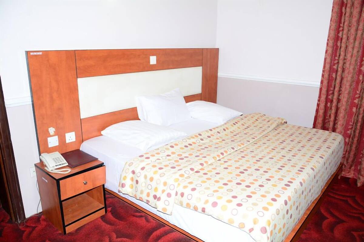 A hotel room with a wooden headboard, white bedding, a patterned bedspread, a bedside telephone, and a red carpet.