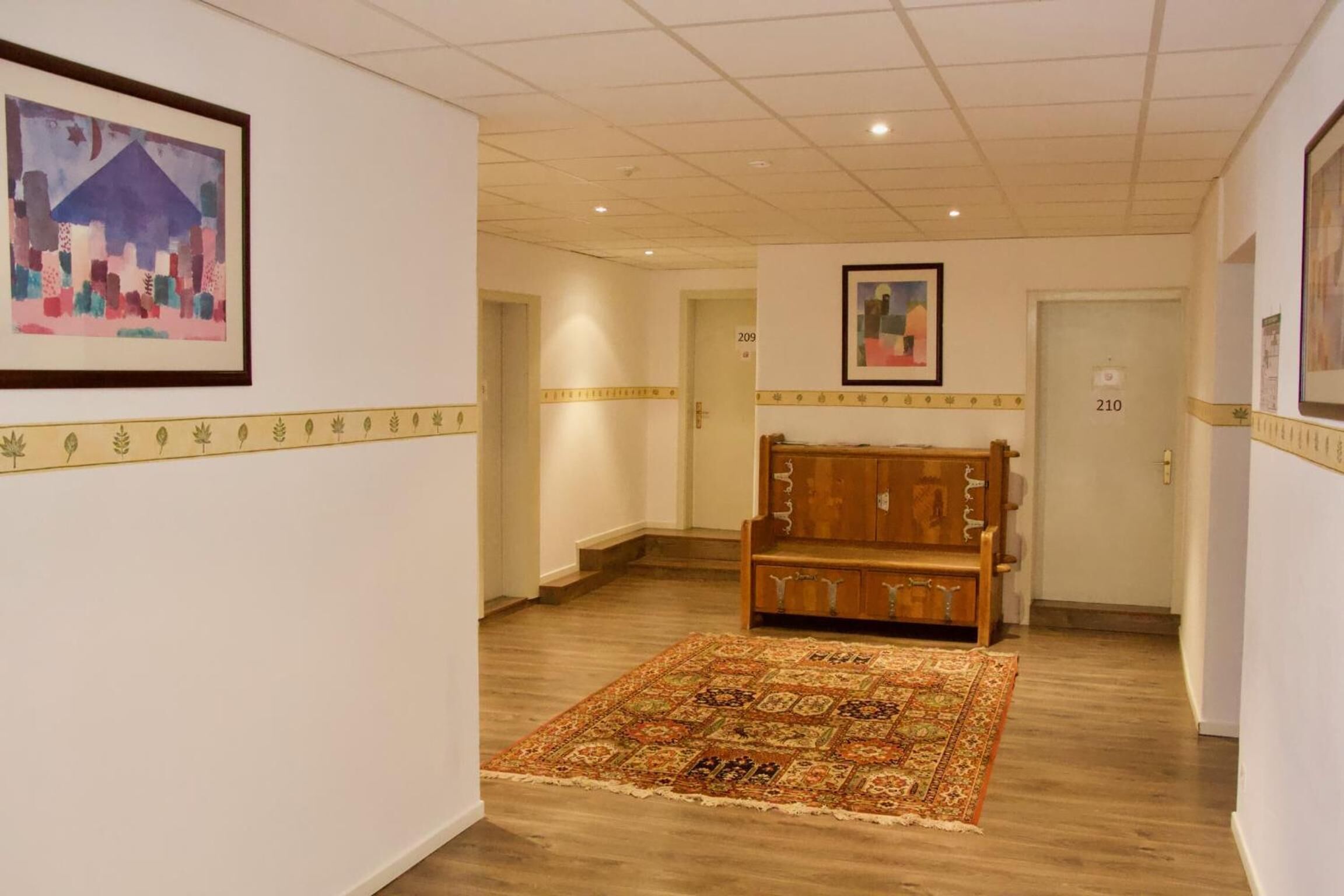 Un couloir d’hôtel avec un tapis à motifs, un banc en bois et des œuvres d’art encadrées accrochées aux murs.