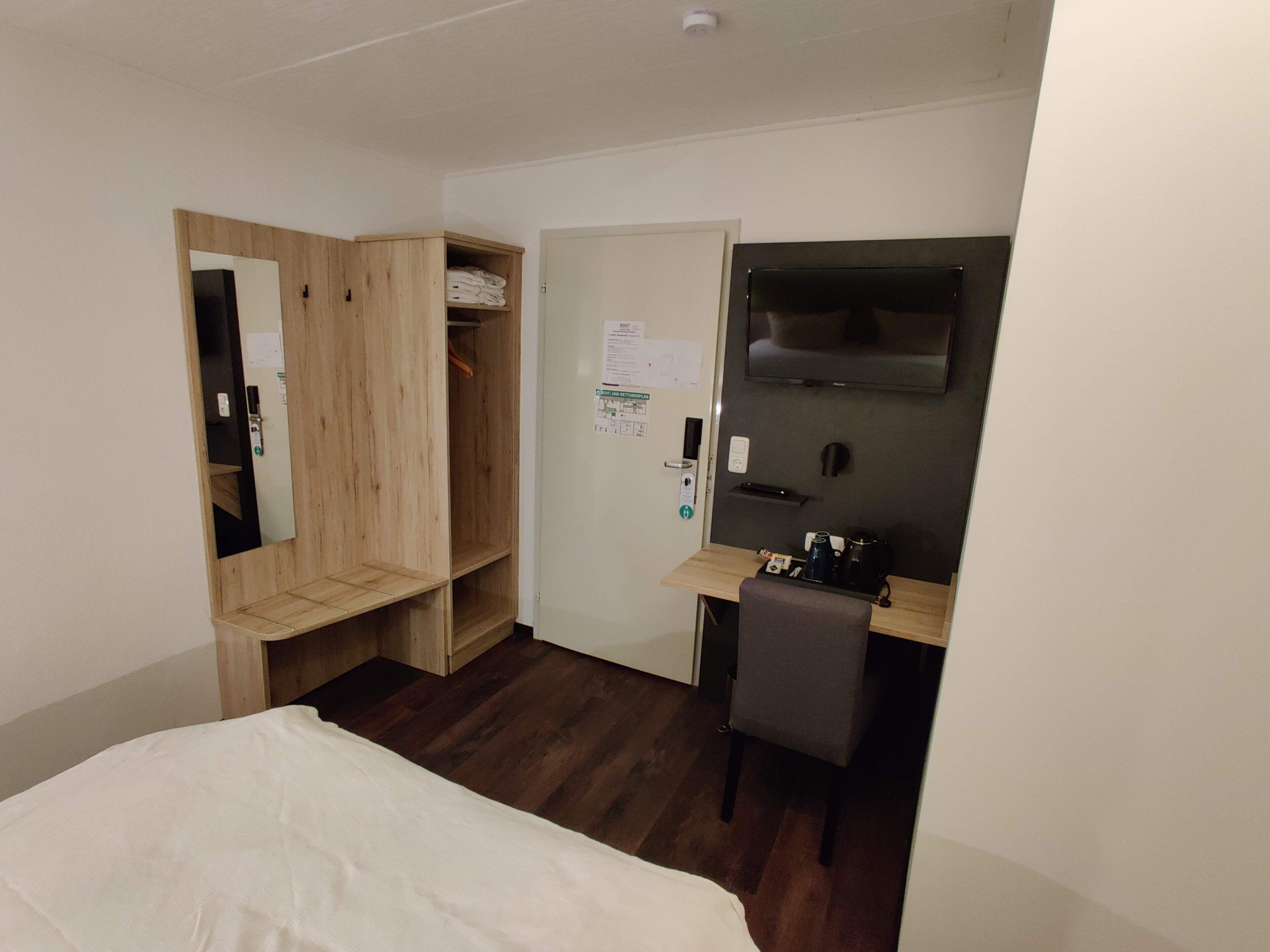 A hotel room with a bed, a wooden wardrobe, a desk with a chair, a flat-screen TV, and a door with a notice.