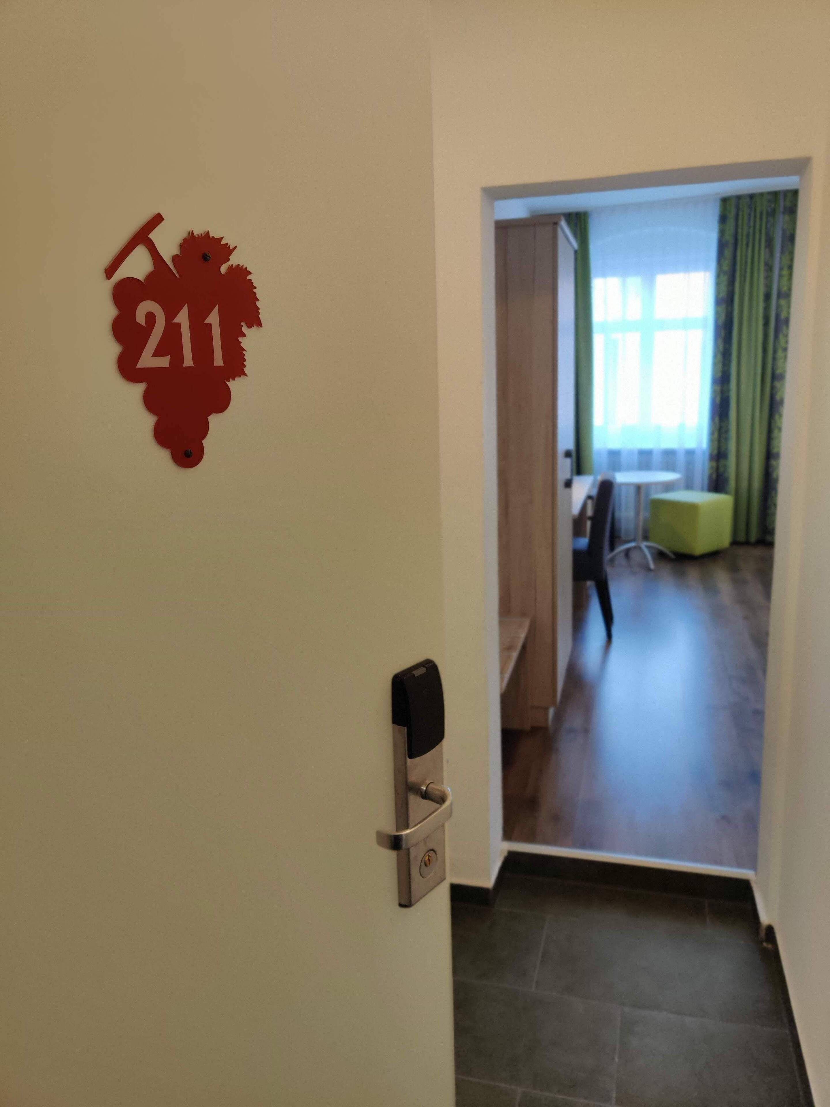 Entrada a una habitación de hotel con un letrero con el número '211' en la pared, una puerta con cerradura de tarjeta y una vista parcial de una habitación con ventana y cortinas verdes.