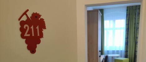 Entrada a una habitación de hotel con un letrero con el número '211' en la pared, una puerta con cerradura de tarjeta y una vista parcial de una habitación con ventana y cortinas verdes.