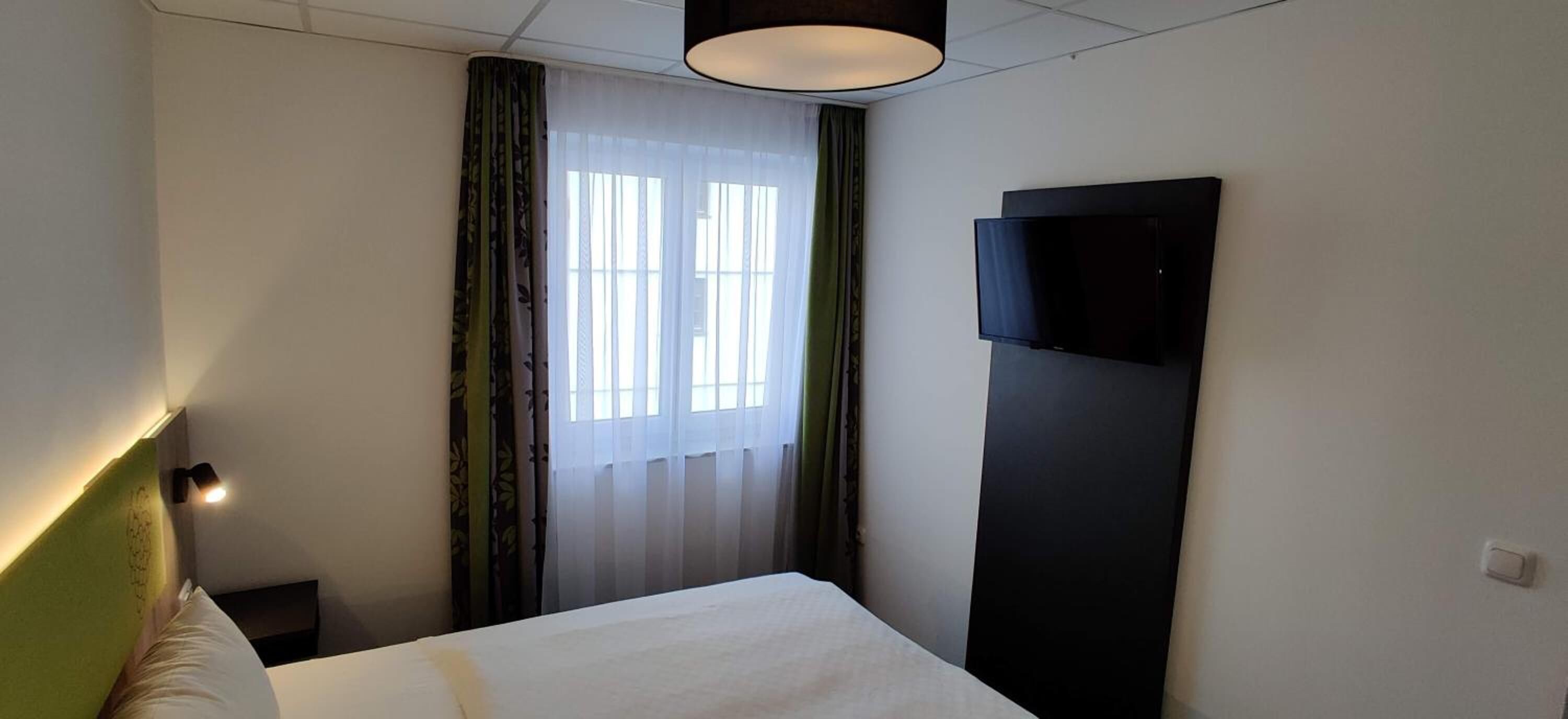 Une chambre d’hôtel avec un lit, un téléviseur fixé au mur, une fenêtre avec des rideaux et une lampe de chevet.