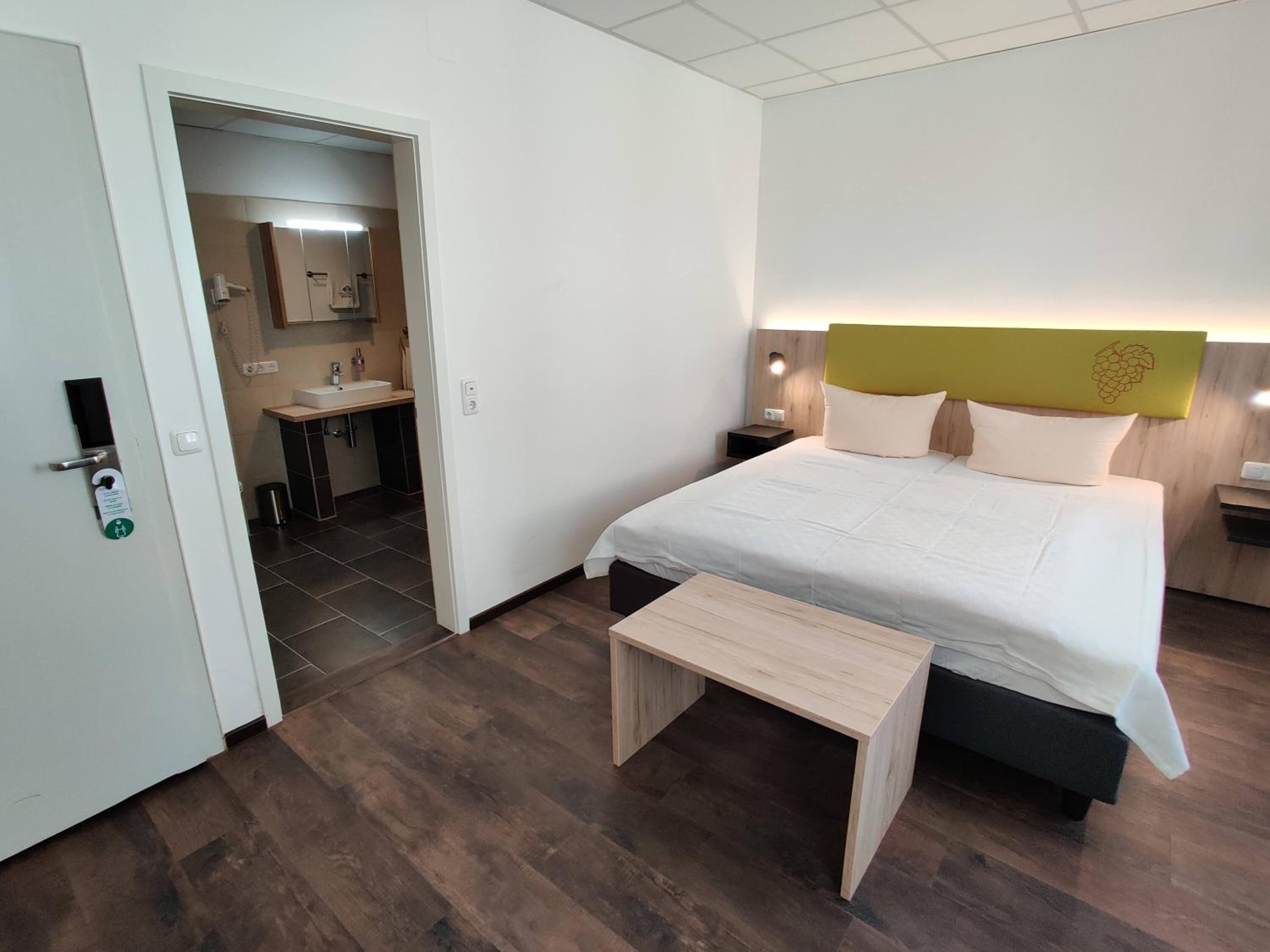 Ein Hotelzimmer mit einem Bett, Nachttischen, einer kleinen Holzbank, einem Badezimmer mit Waschbecken und Spiegel sowie einer Tür mit Kartenlese-Schlösser.