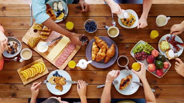 Un grupo de personas comiendo en una mesa de madera con diferentes opciones de desayuno, como pasteles, frutas y bebidas.