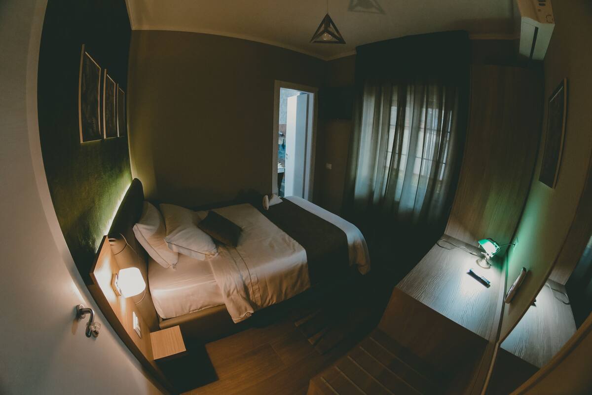 Una camera d'albergo compatta con un letto, un comodino, una lampada e una piccola scrivania con una sedia.