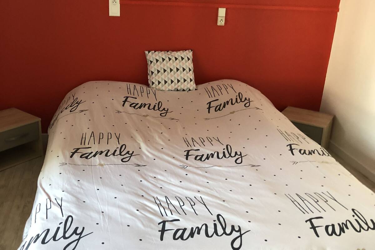 Uma cama com colcha branca com a inscrição 'Happy Family' e um travesseiro geométrico.