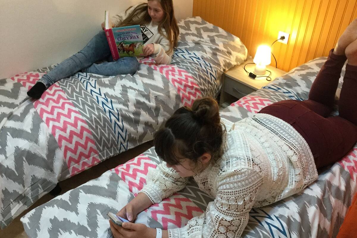 Quarto com duas camas, uma com colcha estampada e outra com colcha lisa. Há uma mesa de cabeceira com luminária e tomada.