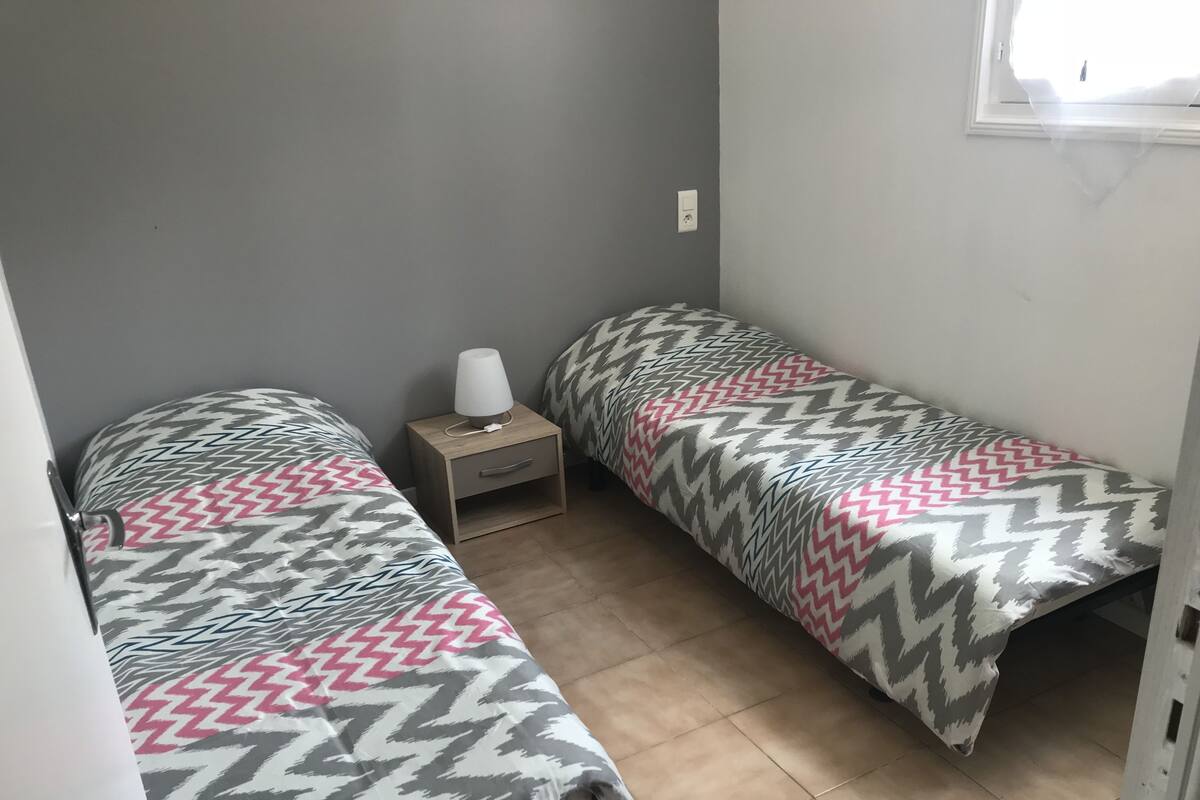 Quarto pequeno e compacto com duas camas de solteiro, cada uma com colcha estampada. Há uma mesinha de cabeceira de madeira com uma luminária entre as camas, e uma gaveta pequena. O quarto tem piso de azulejo e uma janela com moldura branca.