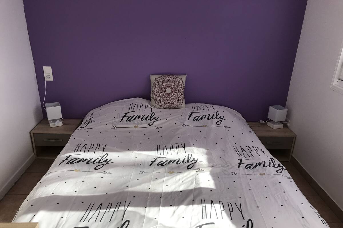 Quarto com parede roxa, cama com colcha temática 'Família Feliz', travesseiro decorativo e mesas de cabeceira com abajures.