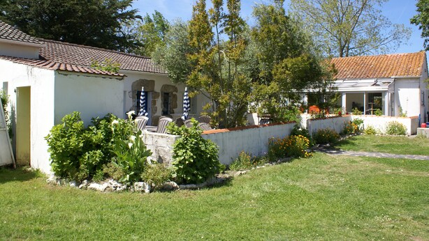A white building with a tiled roof, a stone wall, and a garden with various plants and flowers. - Les Gites De Montmartre (Le Perrier)