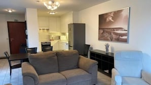 A living room with a sofa, a white armchair, a dining table with chairs, and a kitchen in the background. - Havenzicht 105 (Nieuwpoort)