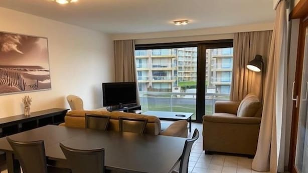 A modern living room with a sofa, a TV, a dining table with chairs, and a view of a residential area through large windows. - Havenzicht 105 (Nieuwpoort)