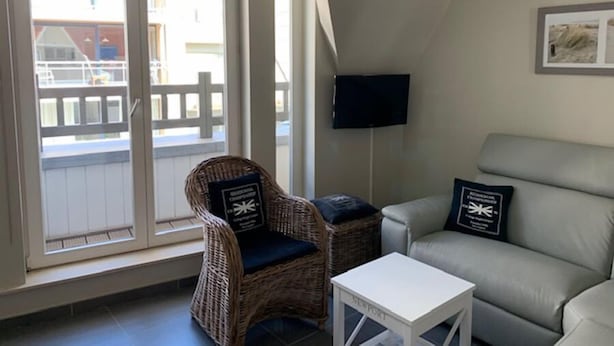 A living room with a wicker chair, a white table, and a grey sofa. There is a television on the wall and a window with a view of buildings outside. - Noorderbries 404 (Nieuwpoort)