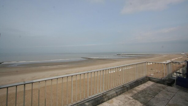 Vista a la playa desde un balcón con barandilla de metal, piso de concreto y un muelle distante.