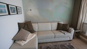 Un salón moderno con un sofá color beige, dos cojines marrones, una mesa de centro de cristal y un gran mural abstracto en la pared.