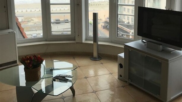 A living room with a large TV, a glass coffee table, and a view of the beach through large windows. - Zeezicht 604 (Nieuwpoort)