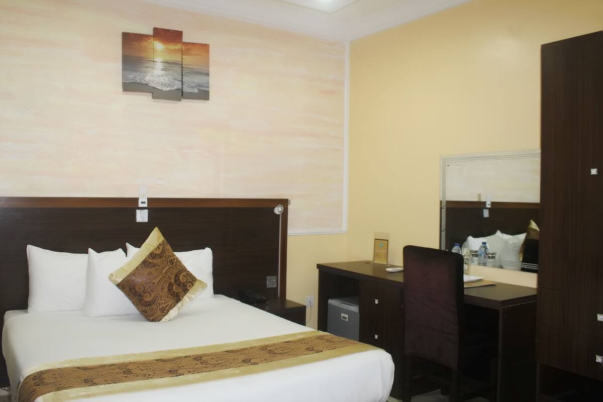 A hotel room with a bed, a desk, a chair, a small trash bin, and a wall-mounted mirror.