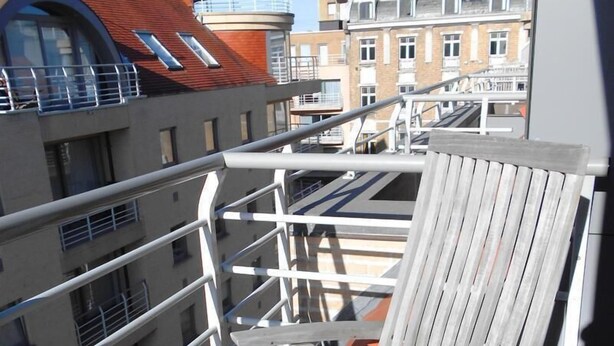 A balcony with a wooden chair and metal railings, overlooking a residential area with buildings and a clear sky. - Cezanne (Nieuwpoort)