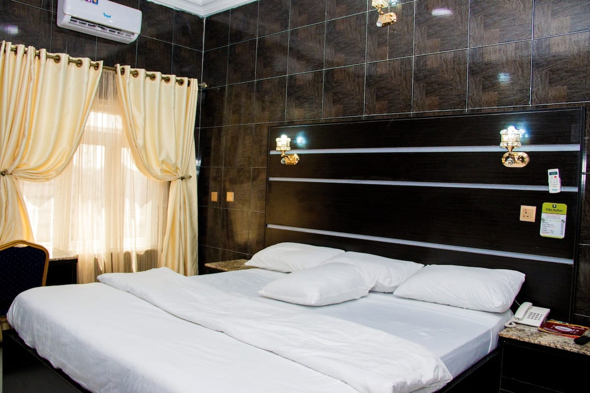 A hotel room with a large bed, a nightstand, a telephone, and a wall-mounted control panel.