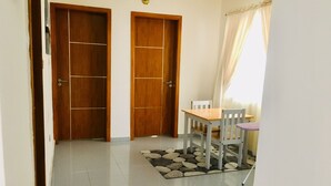 Um corredor com duas portas de madeira, chão de azulejos, uma pequena mesa com cadeiras e uma janela com cortinas.