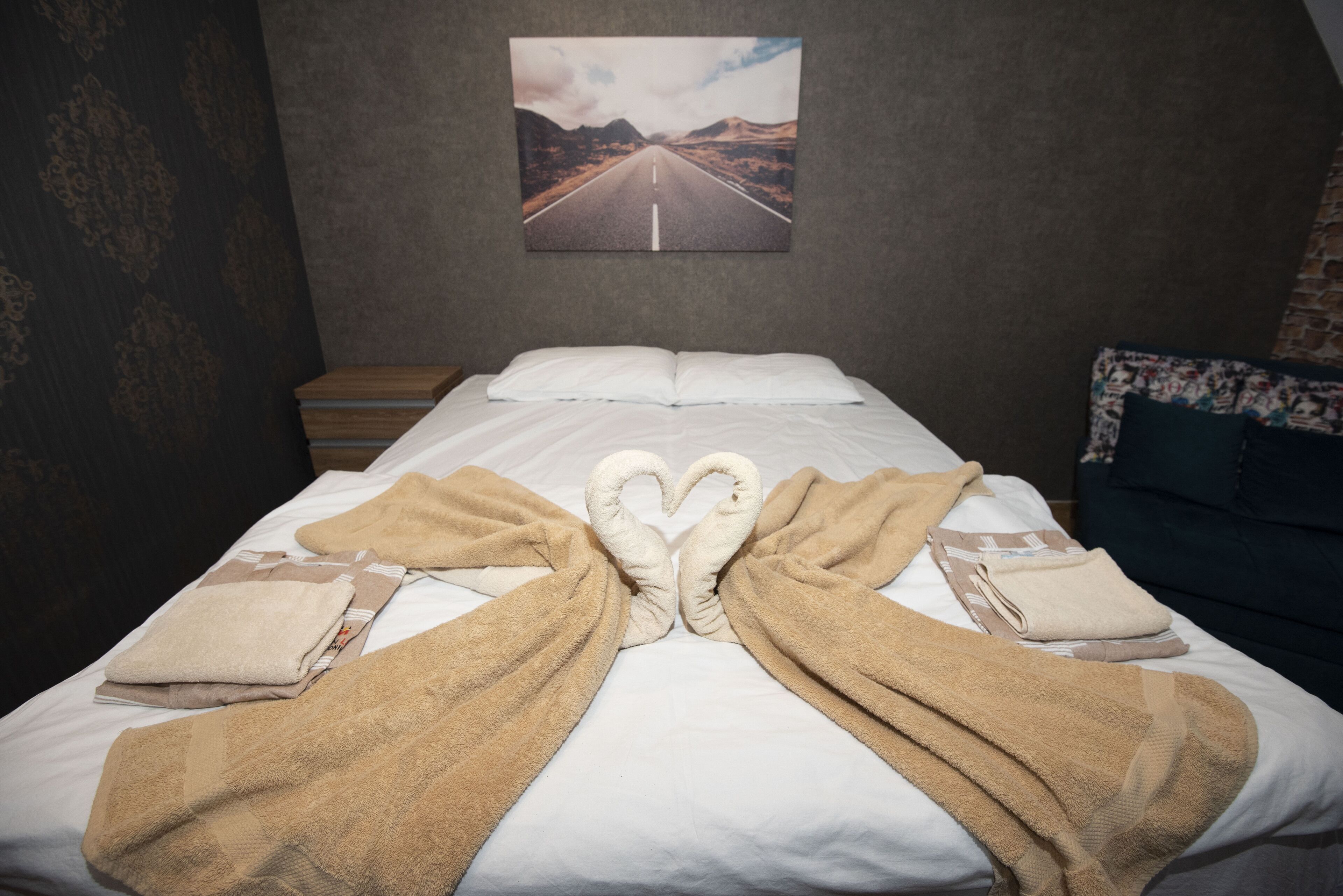 Ein ordentlich bezogenes Bett mit Handtüchern, die zu einem Herz geformt sind, ein Gemälde einer Straße an der Wand und eine Wand mit Muster auf der linken Seite.