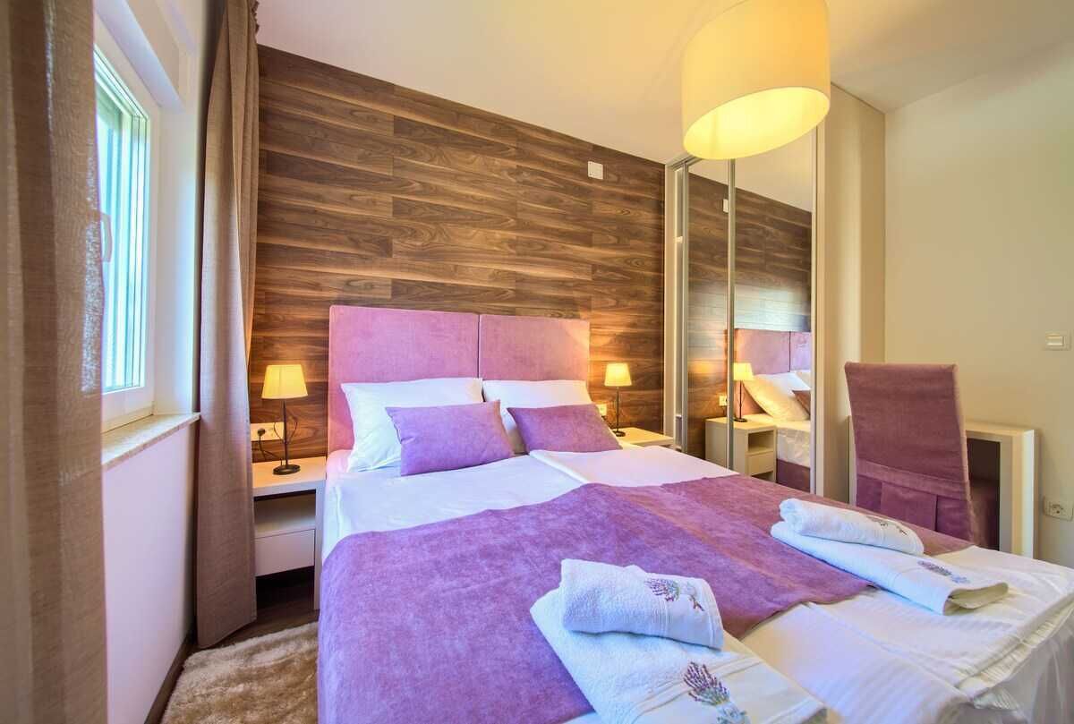 A hotel room with a wooden headboard, a bed with purple bedding, a nightstand with a lamp, a mirror, and a chair with a towel.