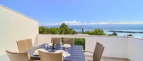 A terrace with a table set for dining, wicker chairs, and a view of a body of water and mountains.