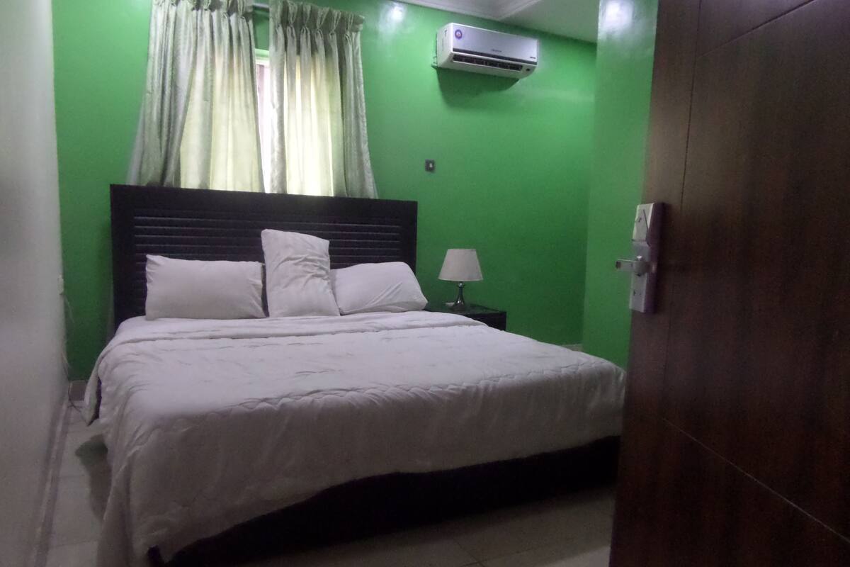 A bedroom with a bed, white bedding, a nightstand with a lamp, a window with curtains, and an air conditioning unit.