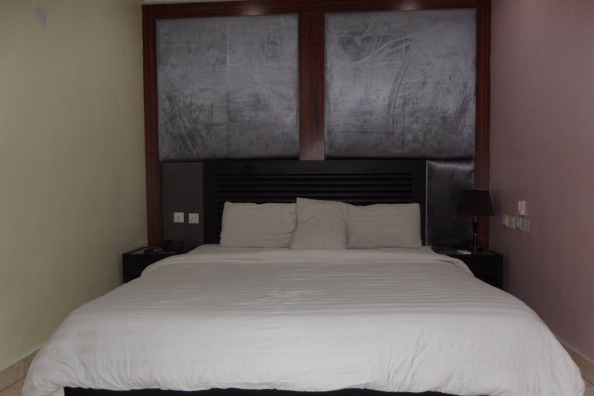 A bedroom with a large bed, a nightstand, a lamp, and a wall-mounted light switch.