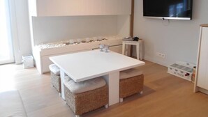 Quarto moderno e bem iluminado com uma mesa branca, cestos de vime, uma pequena mesa de apoio branca, uma TV montada e um armário branco com espaço para armazenamento de vinhos.