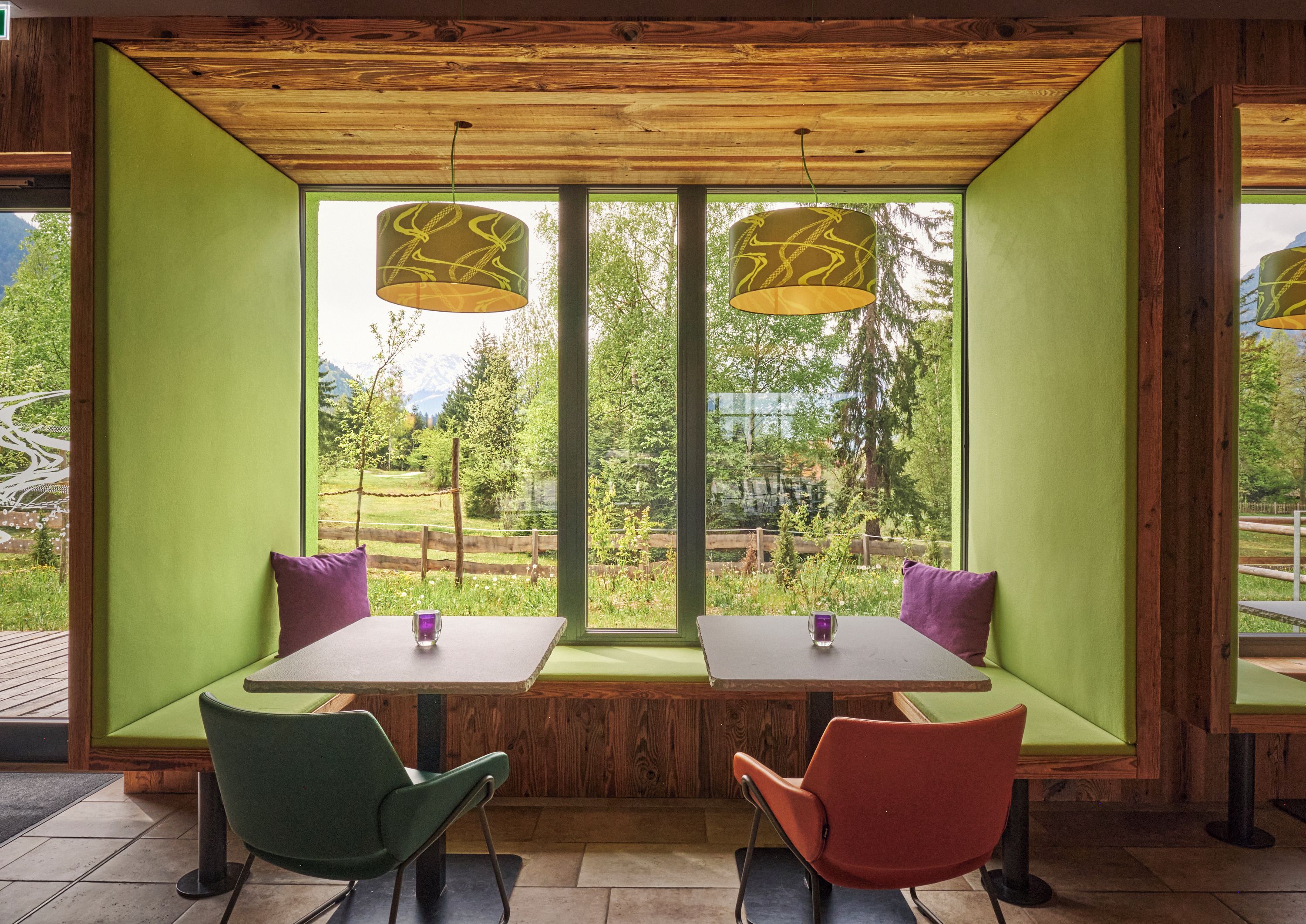 Un comedor con dos mesas y sillas, ventanas amplias con vistas a la vegetación y montañas, e iluminación colgante.