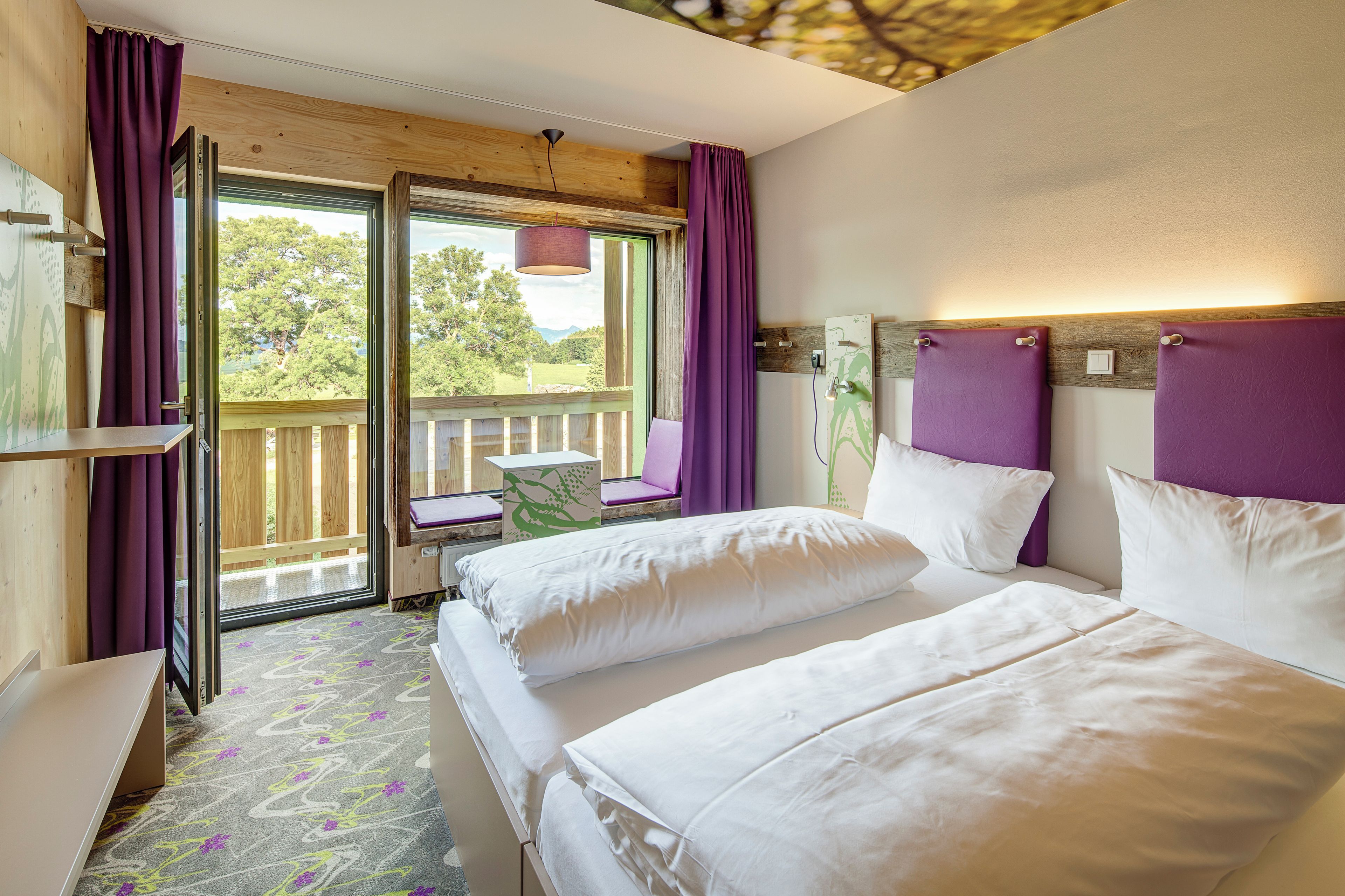 Habitación de hotel con dos camas, un escritorio y una ventana con cortinas moradas.