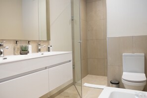 A modern bathroom with a glass shower enclosure, a white toilet, and a vanity with a mirror and two sinks.