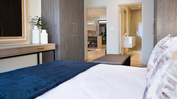 A modern hotel room with a large bed, a desk with two white vases, a mirror, and a view into the kitchen and bathroom. - Hydepark Executive (Randburg)
