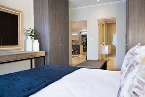Une chambre d’hôtel moderne avec un grand lit, un bureau agrémenté de deux vases blancs, un miroir et une vue sur la cuisine et la salle de bain.