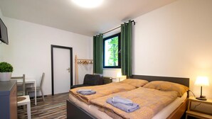 A bedroom with a bed, bedside table, window with curtains, a desk with a chair, and a potted plant. - Penzion Dílna (Trebon)