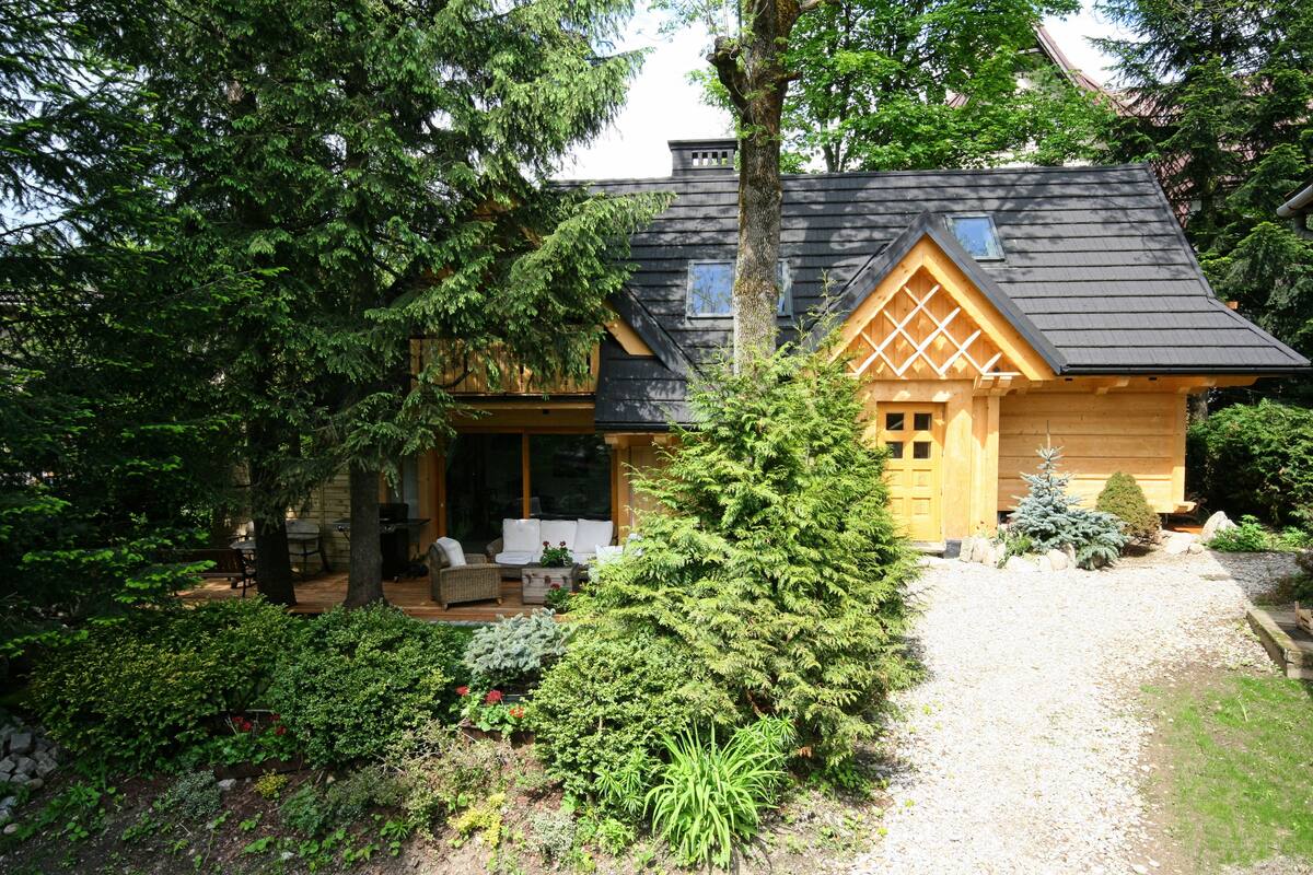 Une cabane en rondins avec un toit en pente, entourée d’une végétation luxuriante et bordée d’une allée en gravier.