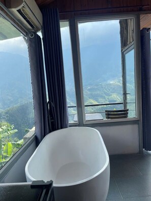 Een badkamer met een bad, een raam met uitzicht op een bergachtig landschap en een klein bankje bij het raam.