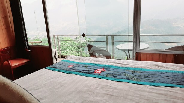 A hotel room with a large bed, a chair, a small table, and a balcony with a view of the mountains. - Valley View Homestay (Sa Pa)