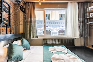 A hotel room with a bed, pillows, a window with curtains, a radiator, and a view of a building outside.