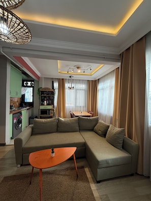 A modern living room with a sectional sofa, a small orange table, and a kitchen area visible through an open door.