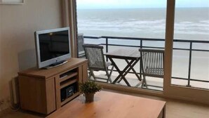 A room with a wooden table, a TV on a wooden stand, and a balcony with a view of the sea. - Scorpio B 902 (Nieuwpoort)