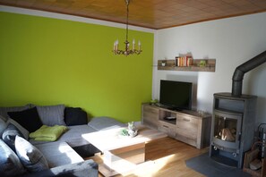 Uma sala de estar com parede verde, sofá cinza, mesa de centro de madeira, televisão sobre suporte de madeira e lareira a lenha.